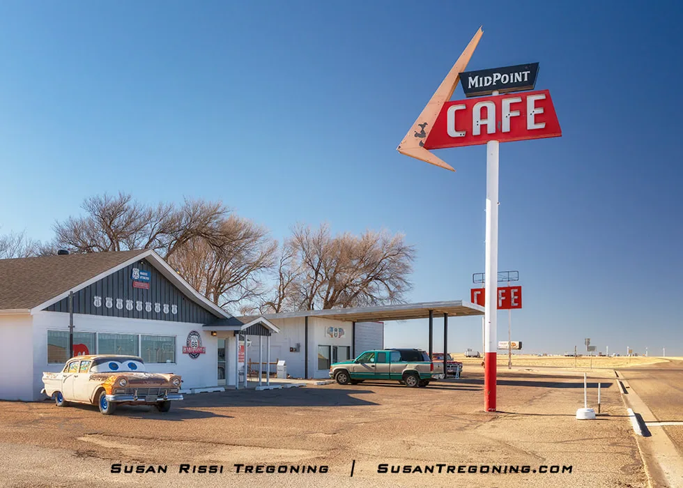 The Midpoint Café on historic Route 66 in Adrian, Texas, with a retro red‑and‑white triangular sign, a low diner building, a white and yellow vintage car with cartoon‑style eyes parked out front, and a green pickup truck nearby under a clear blue sky.