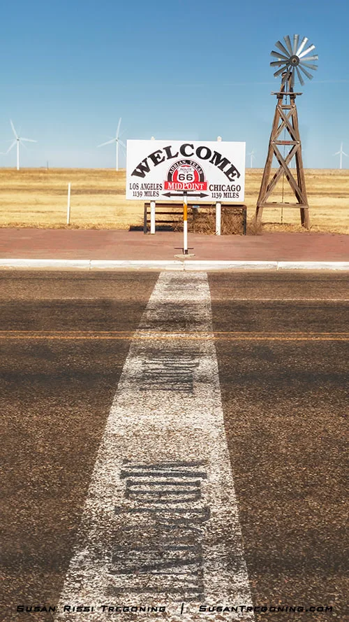 The Route 66 Midpoint line and sign in Adrian, Texas, with a straight highway leading to a white sign that marks the midpoint between Los Angeles and Chicago, a wooden windmill structure behind it, and several wind turbines in the distance under a clear blue sky.