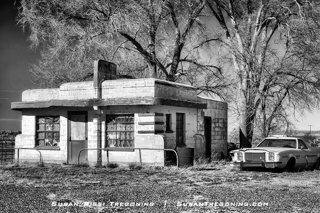 An old Chevrolet El Camino parked beside a weathered concrete‑block building with a flat roof, broken windows, and peeling paint, with metal railing and overgrown grass in front and large leafless trees behind the structure.