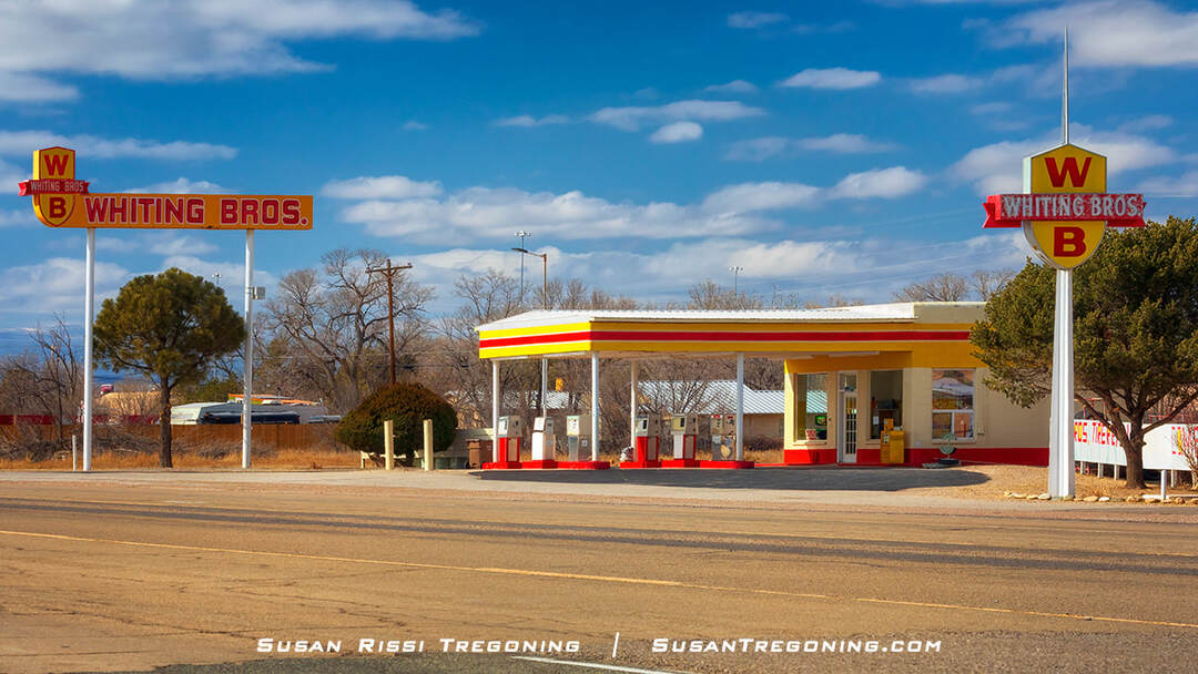 he Whiting Brothers Service Station in Moriarty, New Mexico, with a striped canopy sheltering vintage fuel pumps and two large signs displaying the red and yellow “WB” logo. Trees and shrubs surround the station under a partly cloudy sky.