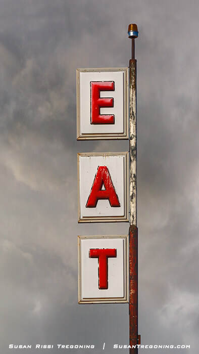 A vertical roadside sign in San Jon, New Mexico, with three stacked panels spelling “EAT” in red letters on a white background. The metal frame shows visible rust, and the sign stands against a cloudy sky.