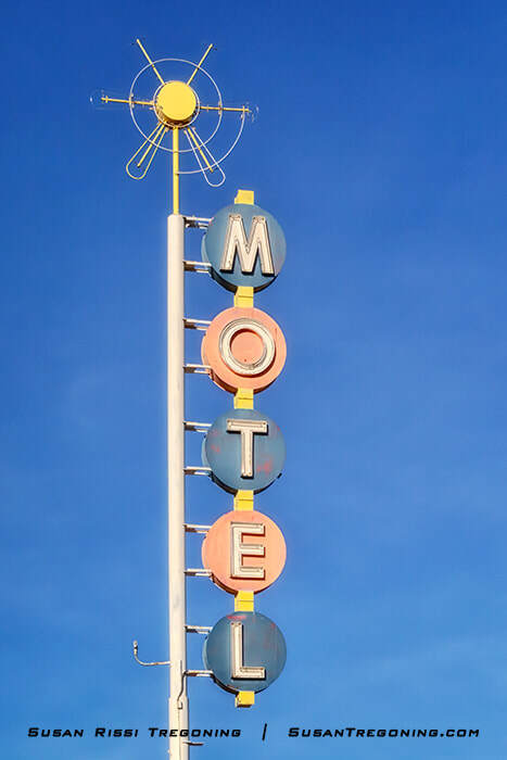 A tall vintage motel sign in Albuquerque with circular blue and pink panels stacked vertically to spell “MOTEL,” topped by a yellow sunburst‑shaped ornament, standing against a clear blue sky.