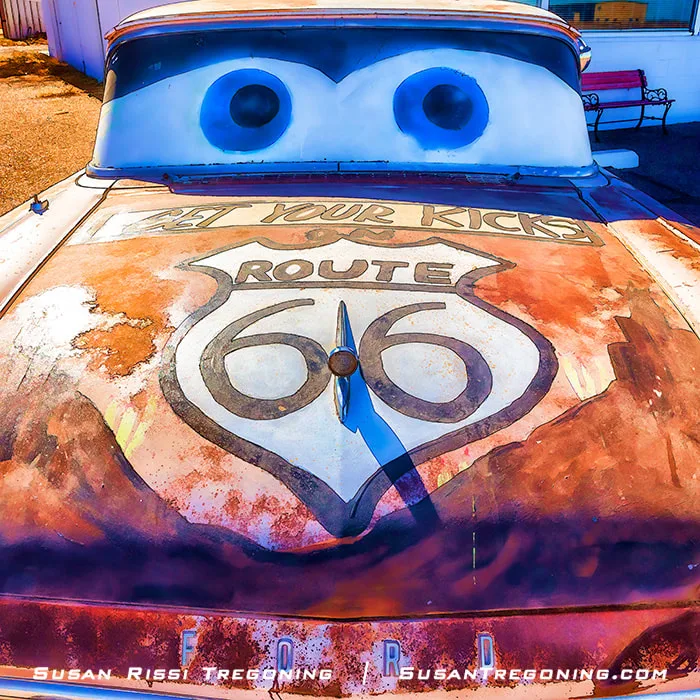 A rusted vintage Ford car painted with a large Route 66 shield and the words “Get Your Kicks!” on the hood, with cartoon‑style eyes painted on the windshield, parked outdoors near a white building and a red bench.
