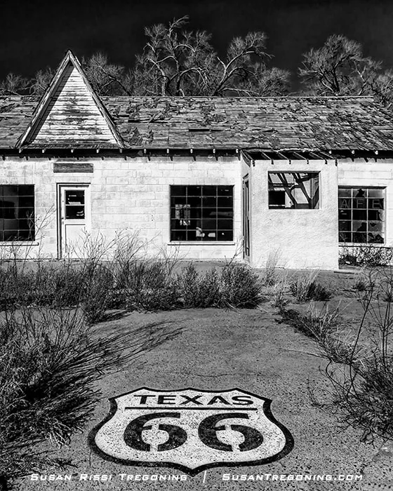 A painted Texas 66 highway shield on the pavement in front of an abandoned stone‑block building with a deteriorating roof, broken windows, and overgrown vegetation, with leafless trees rising behind the structure.