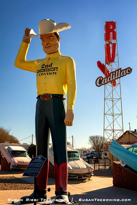 A large roadside statue of the 2nd Amendment Cowboy, dressed in a yellow shirt, blue pants, red boots, and a white hat, tipping his hat beside classic cars and a tall sign structure under a clear blue sky.