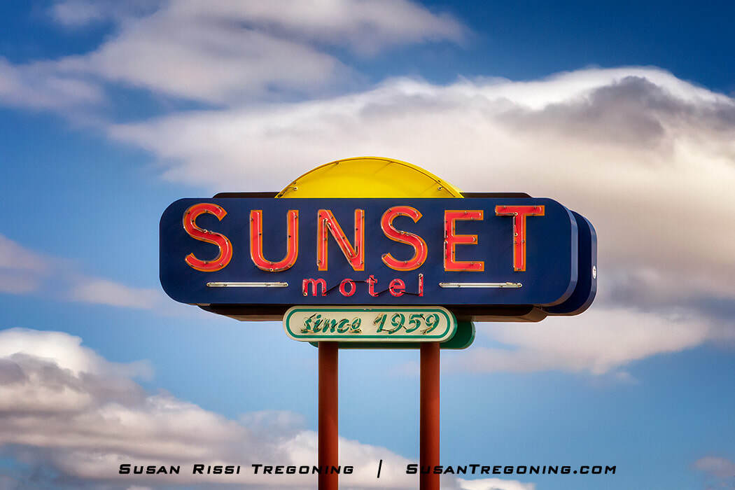 The restored Sunset Motel sign in Moriarty, New Mexico, featuring bold red neon lettering on a blue panel with a yellow semicircle resembling a setting sun above it. A smaller green panel below reads “since 1959,” and the sign stands against a partly cloudy blue sky.