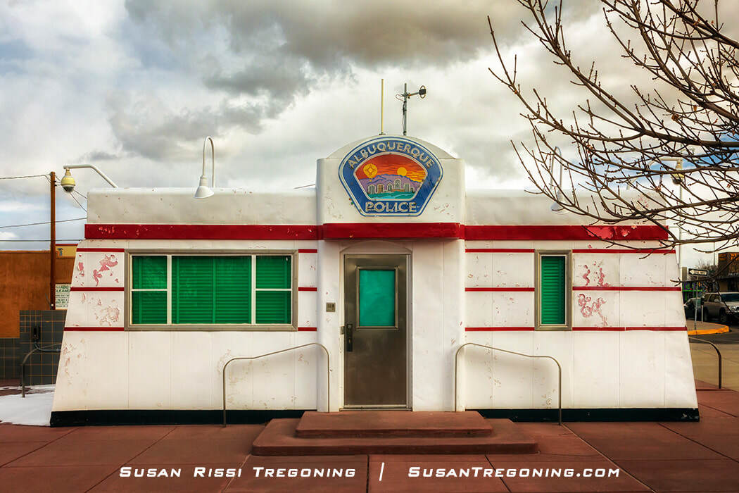 A small prefab building with a retro diner design, featuring a white exterior with horizontal red stripes and green window shutters. A sign above the door reads “Albuquerque Police” with a colorful emblem. The structure sits on a red‑tiled sidewalk with a leafless tree nearby under a cloudy sky.