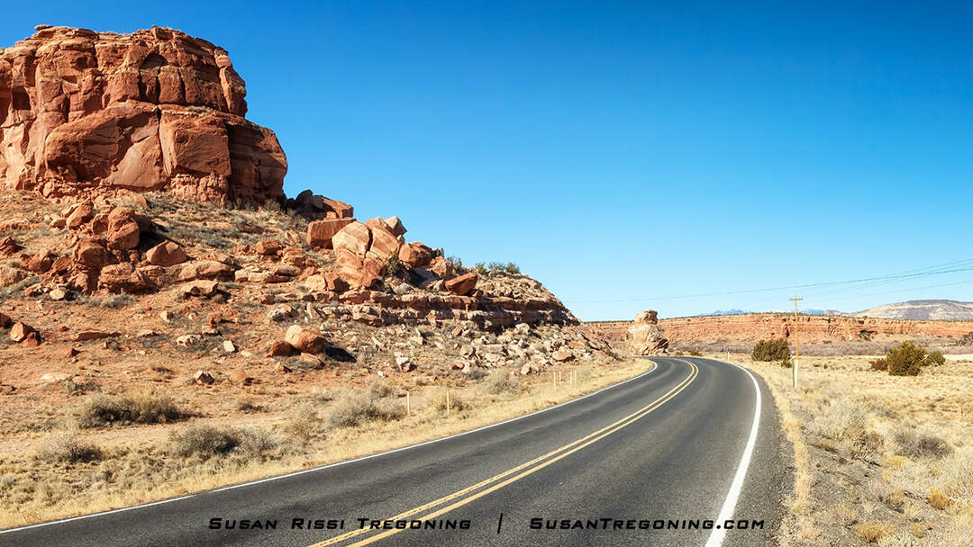A paved road curves through a desert landscape with a large red‑sandstone rock formation on the left. Sparse shrubs and grasses cover the dry terrain, and additional rock formations and mesas are visible in the distance under a clear blue sky.