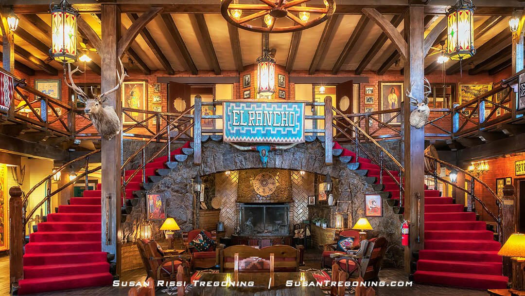 The El Rancho Hotel lobby features an elaborate two‑story interior with a red carpet and a double staircase made of split logs. The staircases curve upward on both sides, with railings formed from naturally bent, polished tree limbs. A large walk‑in stone fireplace sits in a recessed cove beneath the upper‑level balcony that wraps around the room. Heavy dark‑wood furniture, Navajo rugs, and mounted deer heads decorate the space.
