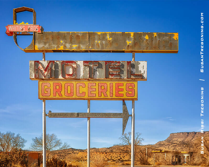 A tall, weathered roadside sign with faded lettering for “Whiting Bros,” “Motel,” and “Groceries” stands in a desert landscape. A large arrow points to the right. Dry vegetation, bare trees, and rocky hills appear in the background under a clear blue sky.