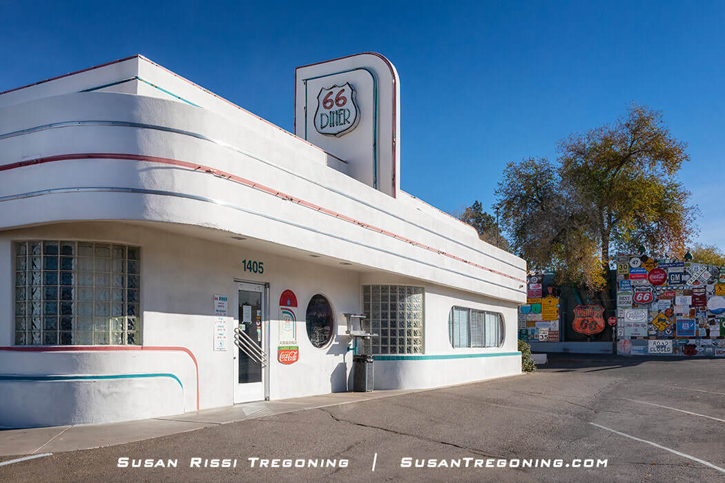 A white Art Deco‑style diner with rounded corners, horizontal colored stripes, and glass block windows. A vertical sign reads “66 Diner,” and the entrance displays the number 1405. A Coca‑Cola logo appears near the door, and a wall covered in vintage road signs is visible in the background under a clear blue sky.