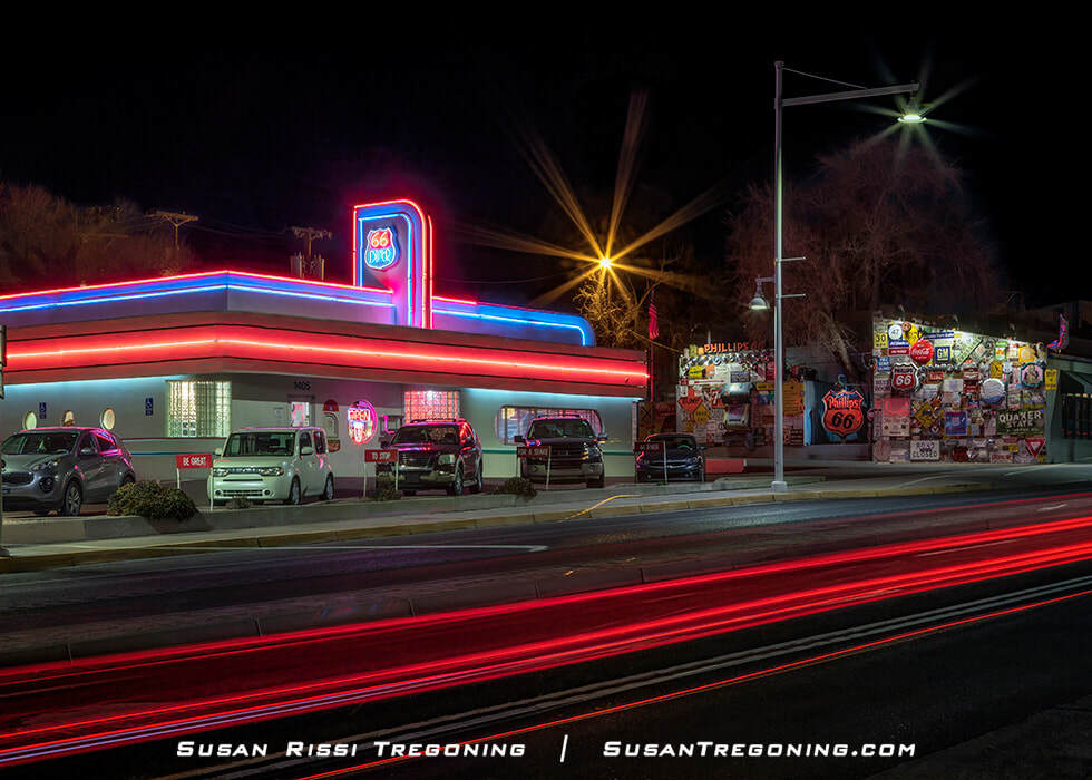 A nighttime view of the 66 Diner with red, blue, and pink neon outlining the building and sign. Cars are parked in front, and red tail‑light streaks from passing vehicles appear across the foreground. A wall covered in vintage signs, including a Phillips 66 sign, is visible across the street under the dark sky.