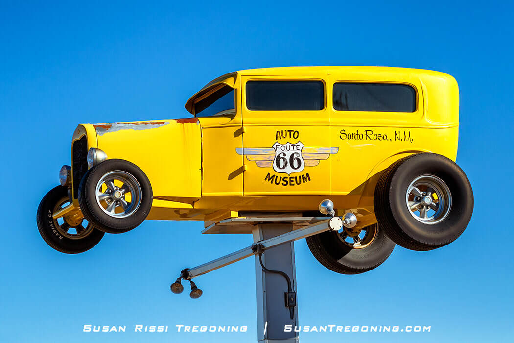 A bright yellow roadster mounted high on a post serves as signage for the Route 66 Auto Museum in Santa Rosa, New Mexico. The car has chrome wheels and visible rust on the front fender, set against a clear blue sky.