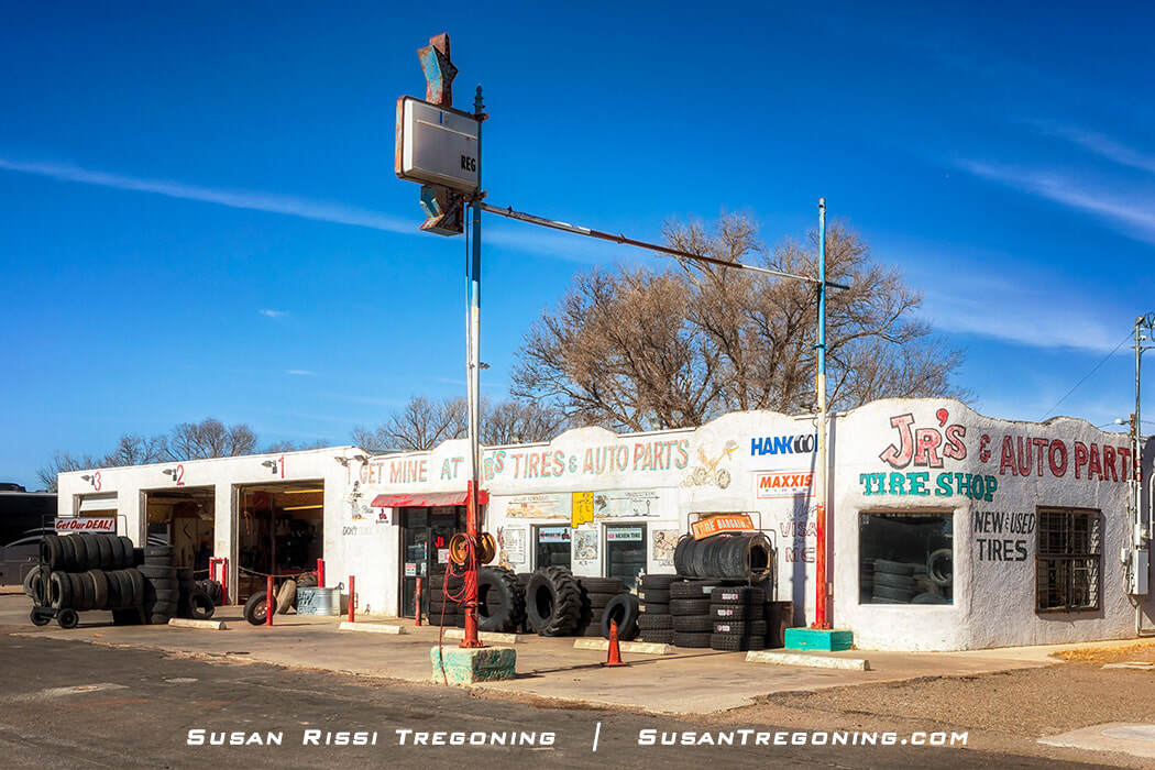 JR’s Tire and Auto Parts in Moriarty, New Mexico, shown as a small stucco building with hand‑painted signage for auto parts and new and used tires. Several stacks of tires sit outside the three open service bays, and an old gas station sign stands above the building. The business occupies the former Greene Evans Garage, an early Route 66 gas station.