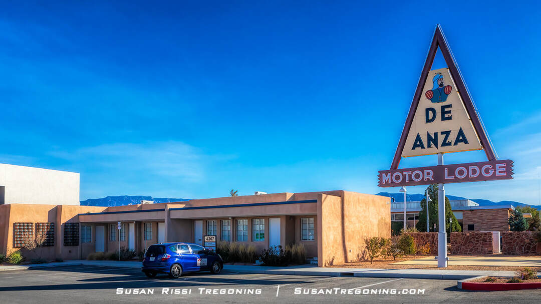 A roadside motel in Albuquerque, New Mexico, with adobe‑style walls and flat roofs. A large triangular sign in front displays an illustrated figure wearing a feathered headdress above the words “De Anza” and “Motor Lodge.” A blue car is parked near the building, and mountains are visible in the background under a clear blue sky.