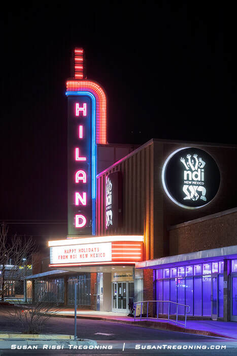 A nighttime view of the Hiland Theater in Albuquerque, New Mexico, showing a tall vertical neon sign reading “HILAND” in pink and blue. Below it, the marquee displays the message “Happy Holidays from NDI New Mexico.” A circular illuminated sign for NDI New Mexico appears on the right side of the building. The entrance area is lit with purple lighting.