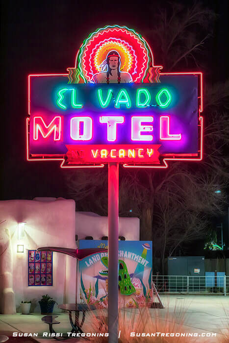 A brightly lit neon sign for the El Vado Motel in Albuquerque, showing a colorful figure in a feathered headdress above the words “El Vado Motel” and “Vacancy.” The sign glows against the night sky, with part of the adobe‑style motel and a mural visible below.