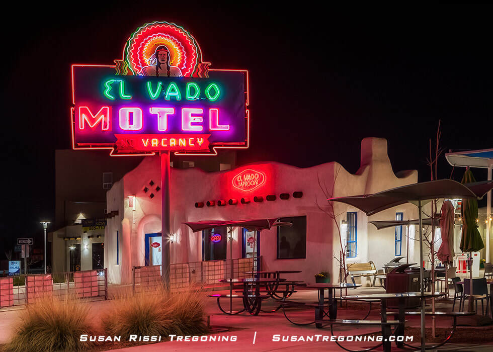 A nighttime view of the El Vado Motel in Albuquerque, New Mexico, showing a brightly lit neon sign with a colorful figure above the words “El Vado Motel Vacancy.” The adobe‑style building behind it features rounded edges and a smaller neon sign reading “El Vado Taproom.” Outdoor tables and umbrellas sit in the foreground under the dark sky.