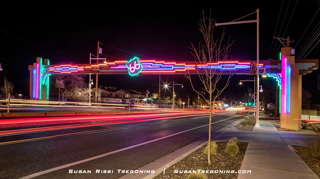 A brightly lit neon arch spans a roadway at night, featuring geometric patterns and a glowing Route 66 shield at the center. Light trails from passing vehicles appear on the street below, with streetlights and buildings visible in the background.