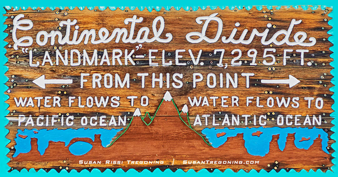 A weathered roadside sign stands beside a paved road in a rural landscape. The sign marks the Continental Divide. Dry vegetation, utility poles, and distant hills appear under a clear blue sky.