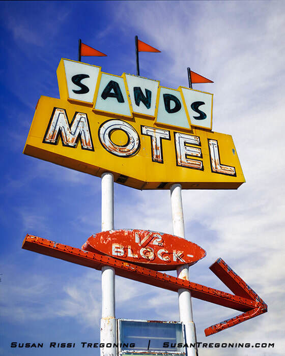 A vintage roadside sign with yellow and blue panels reading “Sands Motel” stands against a blue sky. A red arrow below points to the right with the words “½ Block.” The sign is mounted on two white poles.