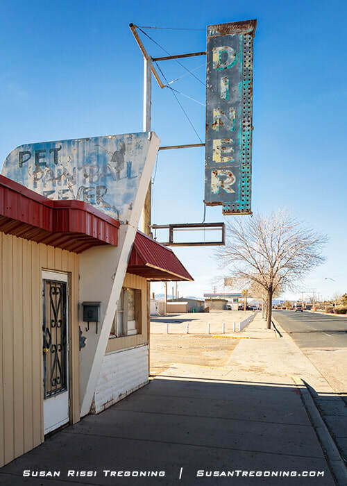 A weathered vertical sign reading “Diner” is mounted above an older building with beige siding and a red awning. The sign shows peeling paint and rust. A faded panel with partially legible lettering is attached to the building. A sidewalk, leafless tree, and clear sky appear in the background.