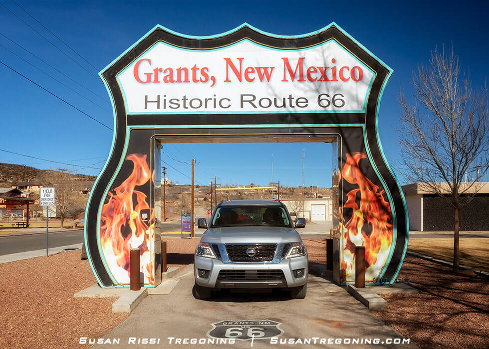 A large roadside arch shaped like a Route 66 shield stands over a paved area. The arch reads “Grants, New Mexico” and “Historic Route 66,” with flame‑style graphics on the sides. A vehicle is parked beneath it, and mountains and utility poles are visible in the background under a clear sky.