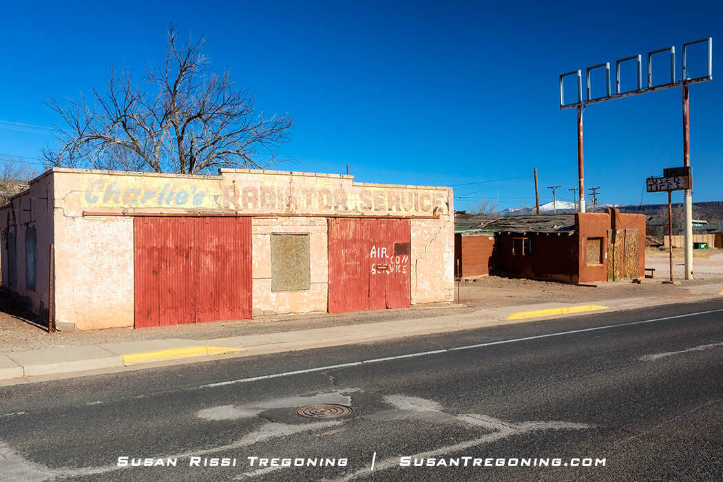 An old building with faded lettering for an automotive service shop stands beside a paved road. The structure has worn paint, two large red doors, and a sign frame above the roof. A bare tree, a smaller building, and distant mountains appear under a clear blue sky.