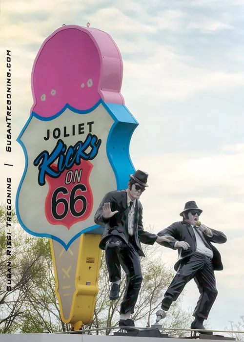 A closeup view shows two statues dressed in black suits, hats, and sunglasses posed in dancing stances beside a large ice‑cream‑cone sign reading Joliet Kicks on Route 66. They stand on the rooftop of the Rich & Creamy building in Joliet, Illinois.