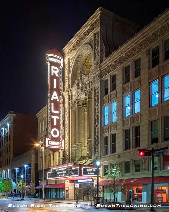 The exterior of the Rubens Rialto Square Theatre in Joliet, Illinois, is shown at night. A tall illuminated vertical sign rises above the entrance, and the marquee below is lit. The façade features classical columns, sculpted figures, and decorative carvings. Streetlights and nearby buildings line the roadway in front.