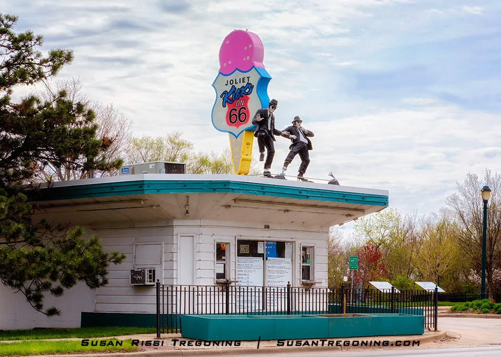 A small roadside building with a teal roof stands among trees and fencing. Two statues resembling the Blues Brothers, dressed in black suits, hats, and sunglasses, appear in dancing poses atop the roof. Above them is a large ice‑cream‑cone sign reading Kicks on 66. The structure is part of Route 66 Park in Joliet, Illinois.