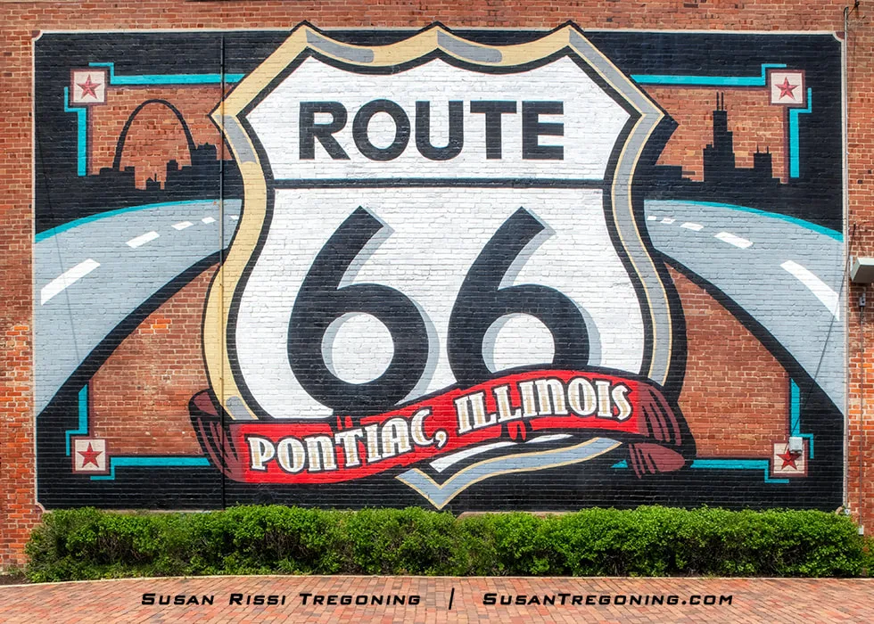 A large mural painted on a brick wall features a Route 66 shield above a red banner reading Pontiac, Illinois. A curved road graphic leads toward stylized skylines on each side. Red bricks cover the ground in front of the mural; these are original Route 66 bricks designed to be driven on. The mural sits behind the Illinois Route 66 Hall of Fame Museum Complex.