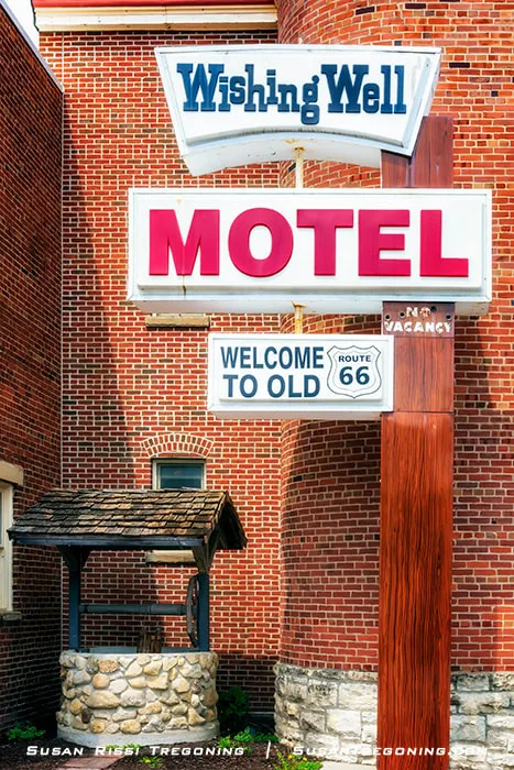 A vintage roadside sign reading Wishing Well Motel stands beside a stone wishing well with a wooden roof. The sign includes additional panels referencing Old Route 66 and a no‑vacancy notice. A brick building appears in the background. These artifacts were relocated from the former Wishing Well Motel in Countryside, Illinois, and are now displayed behind the Illinois Route 66 Hall of Fame Museum Complex.
