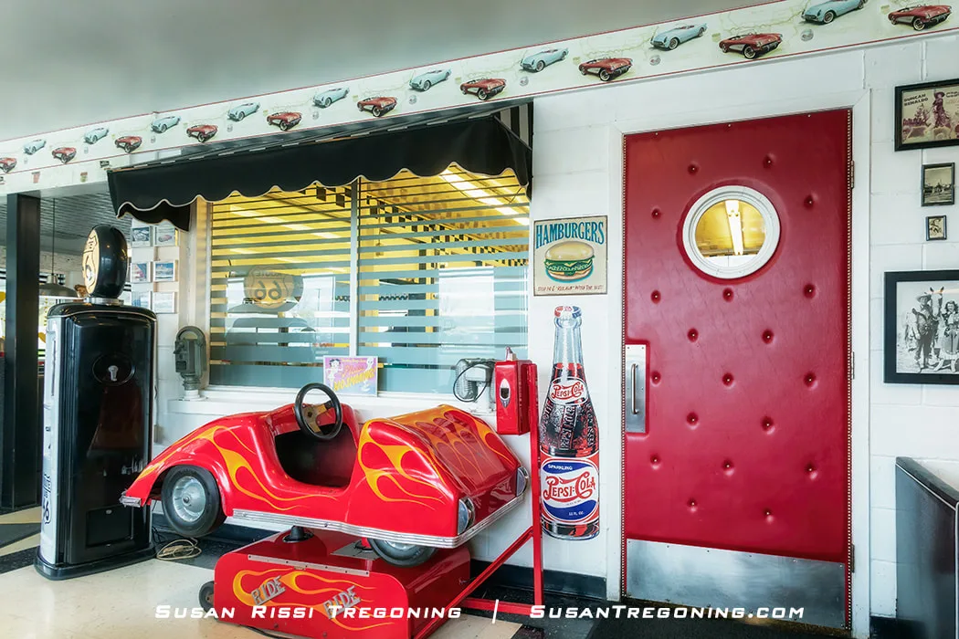 A red coin‑operated race‑car kiddie ride sits inside the Polk‑A‑Dot Drive‑In. The car has yellow flame graphics and is positioned near a red padded door with a round window. A wall‑mounted payphone, vintage posters, a black gas pump, and a black‑and‑white checkered floor contribute to the retro diner setting.
