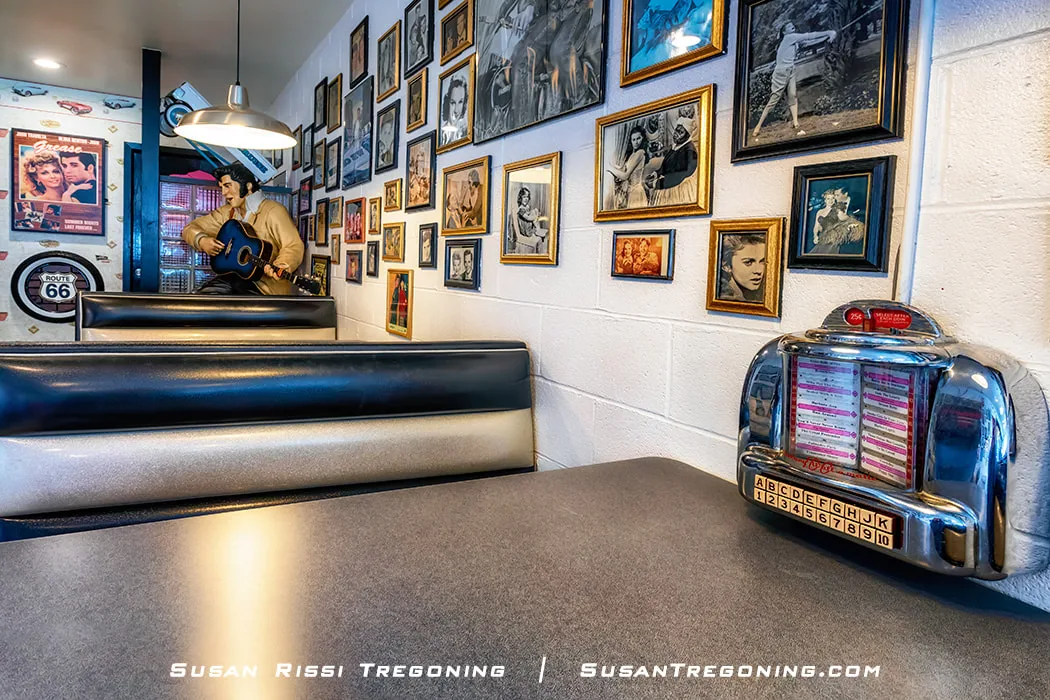 A retro diner booth with black and silver seating sits against a white brick wall covered in framed photographs and portraits. A small tabletop jukebox is mounted at the end of the booth. A life‑sized statue of a musician holding a guitar stands in the background, and a Route 66 sign and vintage posters add to the mid‑century décor.