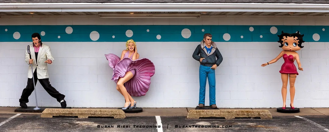 Four life‑sized statues stand in a row outside the Polk‑A‑Dot Drive‑In. From left to right are figures resembling Elvis Presley holding a microphone, Marilyn Monroe in a dress‑blown pose, James Dean in a leather jacket with his hands in his pockets, and Betty Boop in a red dress. They are positioned in front of a white wall with a turquoise stripe and circular accents, behind concrete parking barriers.