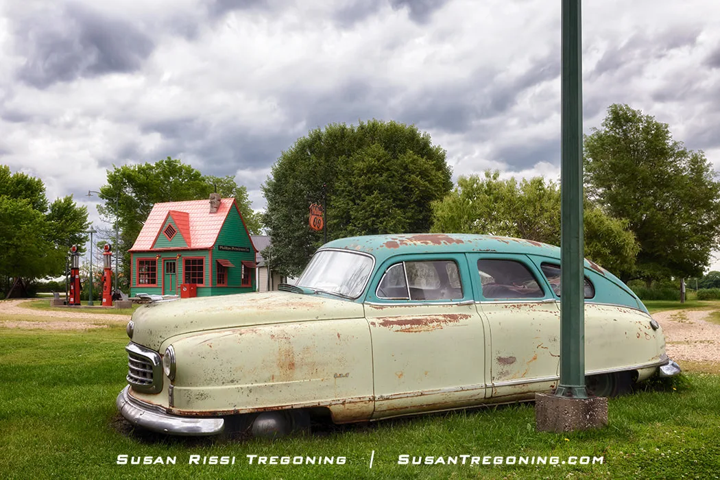 A weathered 1949 Nash Airflyte 600 sits on grass in front of a restored 1920s cottage‑style Phillips 66 service station at Red Oak II, with two red vintage gas pumps and a green‑and‑red building framed by overcast skies and surrounding trees.