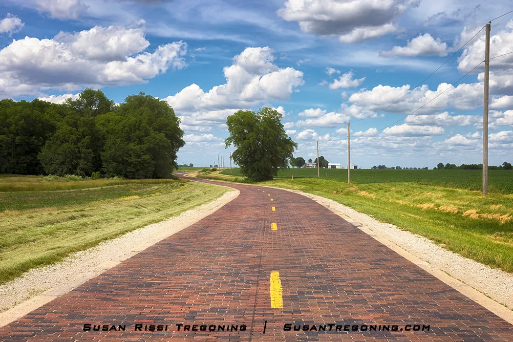 The historic Auburn Brick Road in Illinois is shown as a restored 1.53‑mile hand‑laid brick segment of the original 1926–1930 alignment of U.S. Route 66. The roadway curves through a rural landscape, with the brick surface visible along its length.