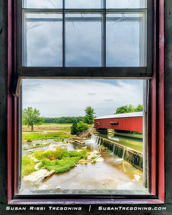 A red covered bridge is seen through the open window of the historic Bridgeton Mill, framed by the red window trim, with a small waterfall, stream, rocks, and trees visible outside under a partly cloudy sky.