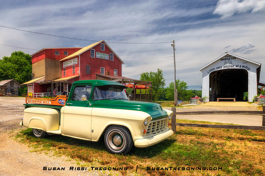   
A restored two‑tone 1955 Chevrolet 3100 pickup truck is parked in front of the red Bridgeton Mill, with the white Bridgeton Covered Bridge visible to the right, surrounded by trees under a partly cloudy sky.