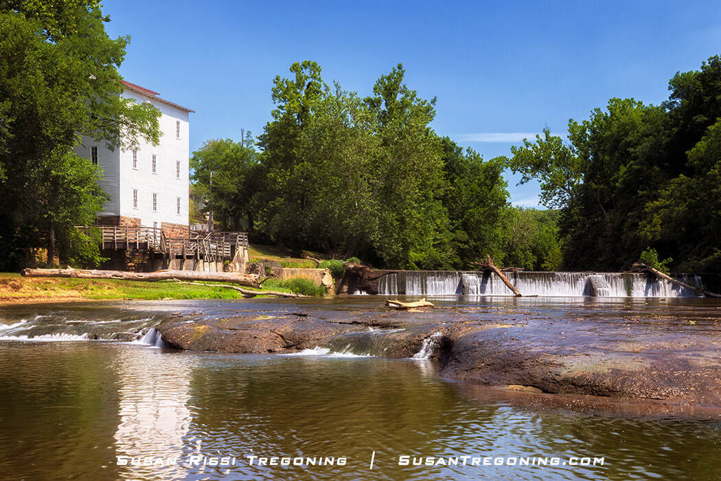 The Mansfield Roller Mill stands beside Big Raccoon Creek, a large white wooden mill building overlooking the water near a small waterfall. Rocks and ripples fill the foreground of the creek, with dense green trees surrounding the mill under a clear blue sky.