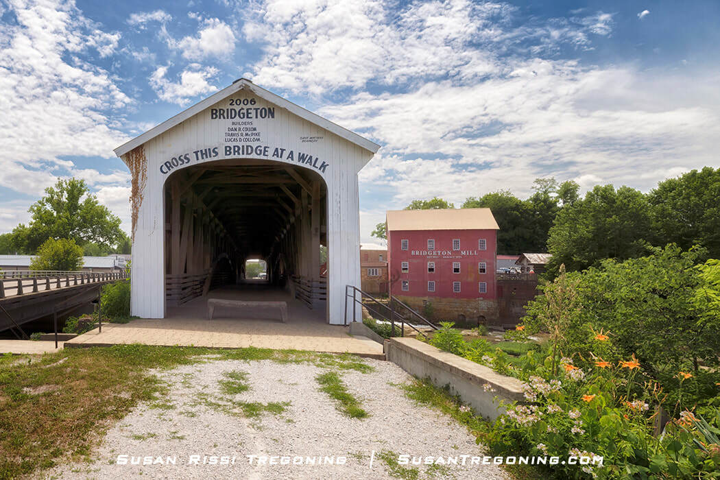 The white‑painted Bridgeton Covered Bridge, rebuilt in 2006, spans Big Raccoon Creek with the red Bridgeton Mill visible behind it. The bridge crosses a small waterway toward the mill, surrounded by green trees under a partly cloudy sky.
