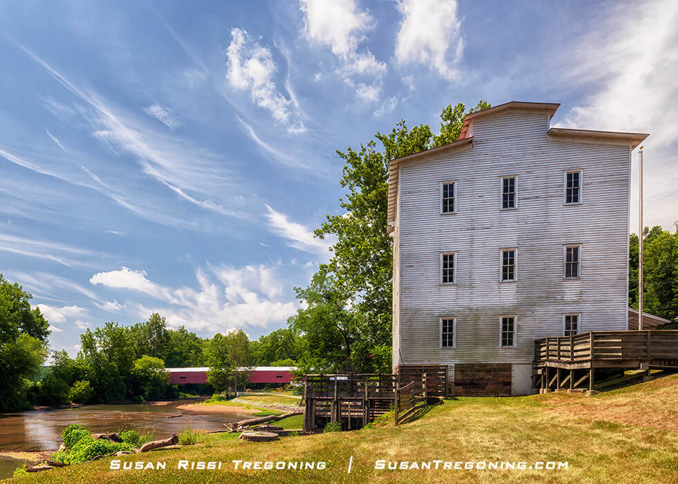 The historic Mansfield Roller Mill stands beside Big Raccoon Creek, a large white wooden mill building with multiple windows overlooking the water. Just downstream, the red Mansfield Covered Bridge spans the creek, surrounded by green trees under a bright blue sky with wispy clouds.