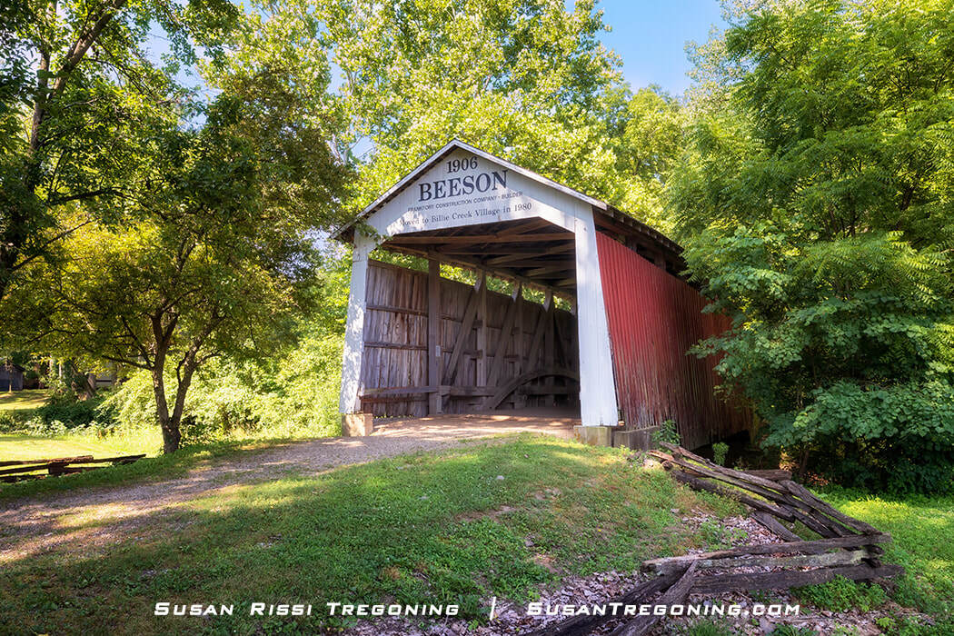 Beeson Covered Bridge, a white‑front, red‑sided wooden bridge with a peaked roof, set among green trees and grass on a sunny day.
