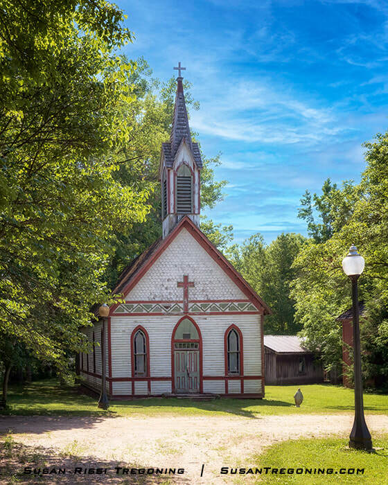 The 1886 St. Joseph’s Catholic Church, a white wooden structure with a tall steeple and arched windows, standing among green trees at Billie Creek Village after being moved from Rockville, Indiana.