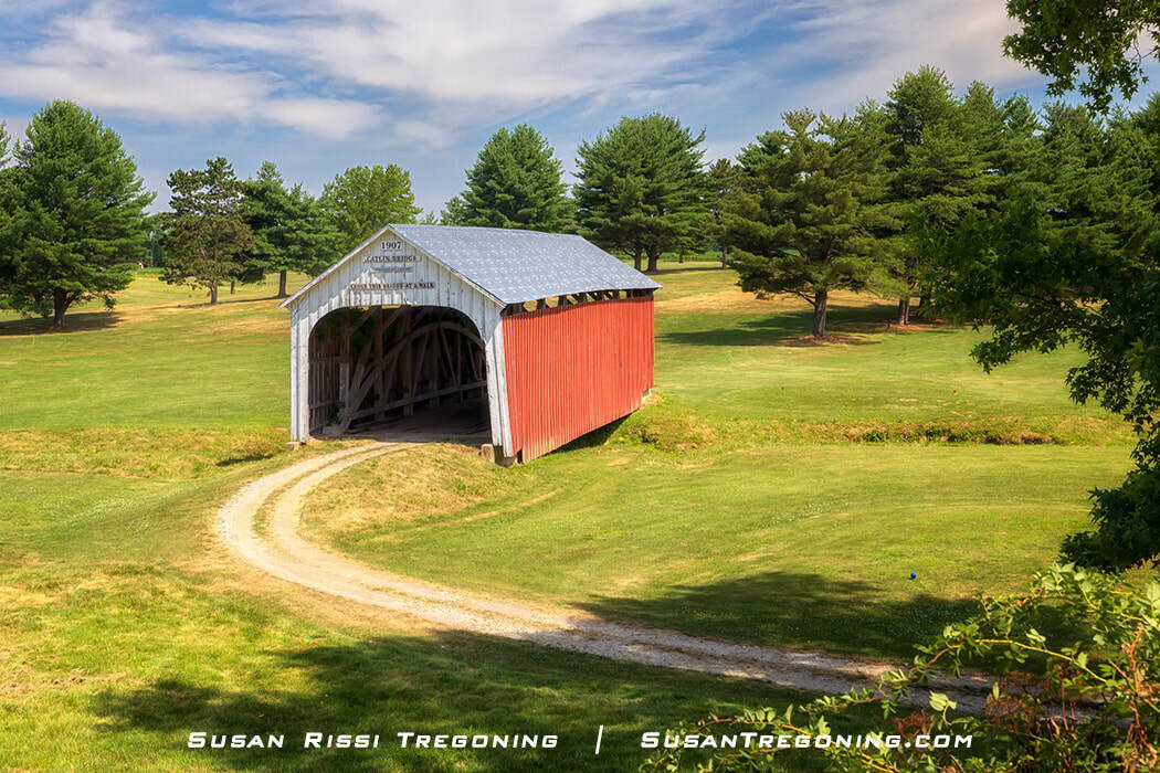 Catlin Covered Bridge, a red wooden covered bridge with white trim and a metal roof, set along a curving dirt path at the Parke County Golf Course in Rockville, Indiana, surrounded by trees and open grassy areas.