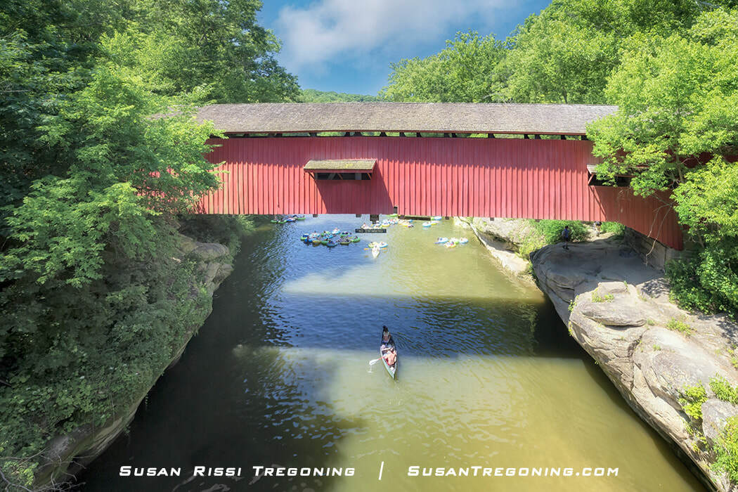   
People float down Sugar Creek on inner tubes and paddle a canoe beneath the red Narrows Covered Bridge, surrounded by lush green trees and calm summer water.