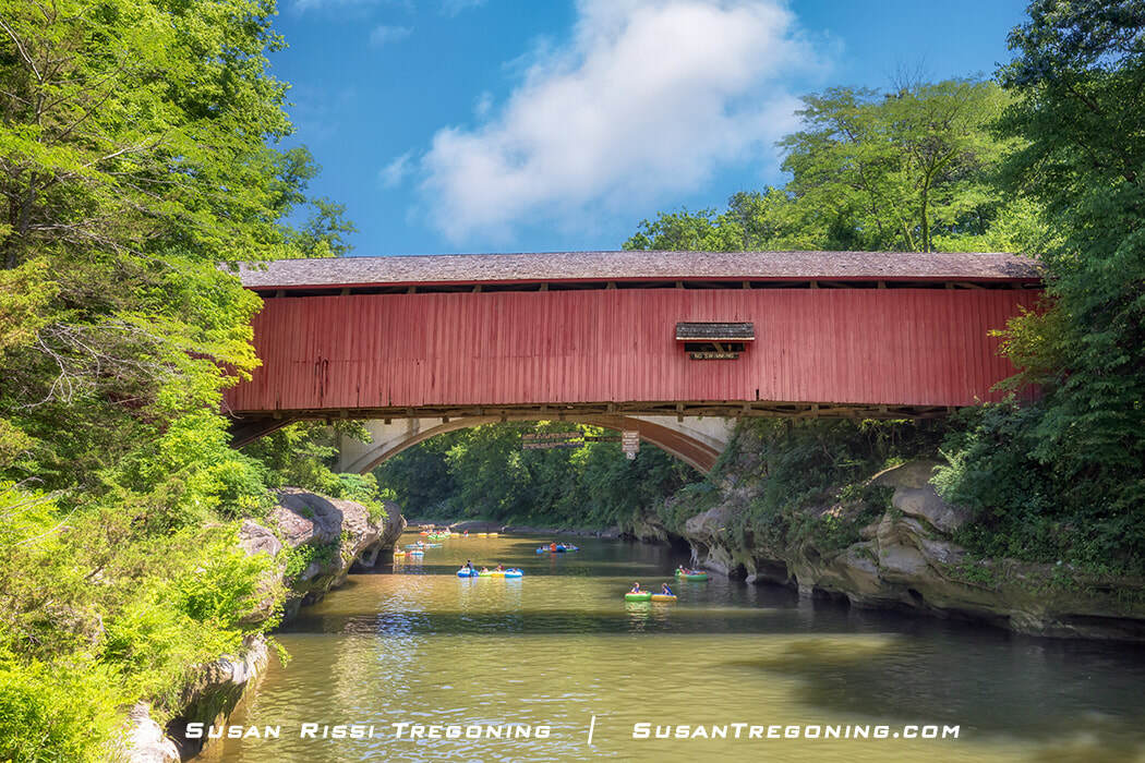   
People float down Sugar Creek in kayaks and inner tubes beneath the red Narrows Covered Bridge, surrounded by lush green trees and calm summer water.
