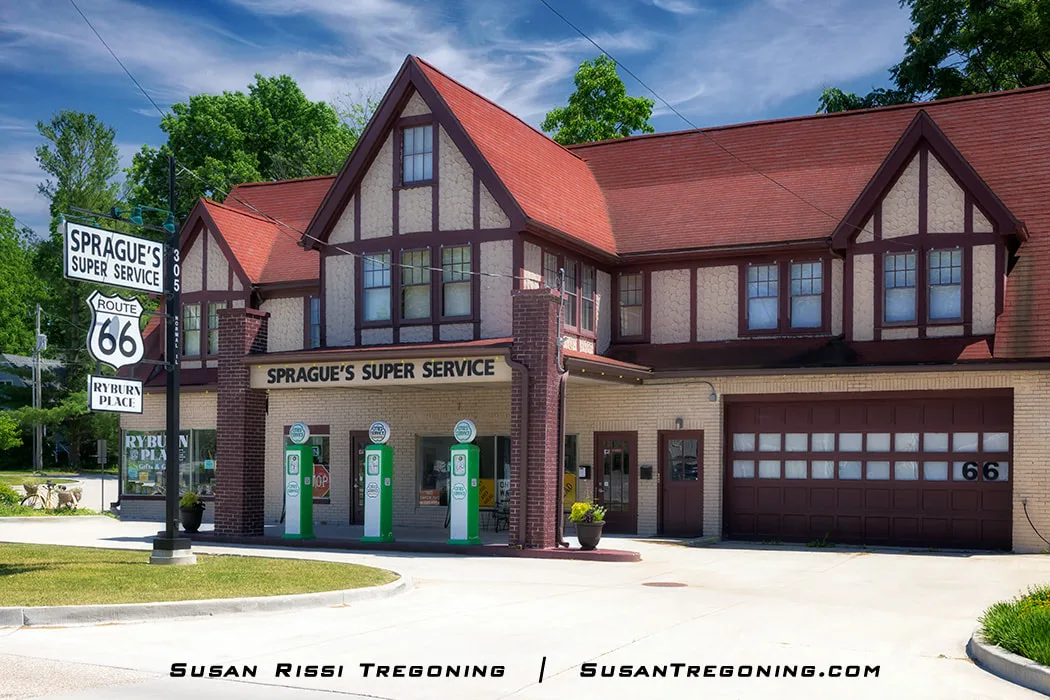 A Tudor Revival service station with a steep red roof and beige brick walls stands along Route 66 in Normal, Illinois. A canopy shelters three vintage green and white gas pumps, and a vertical sign identifies the site as Sprague’s Super Service. The building is two stories tall and features a garage door marked with the number 66.