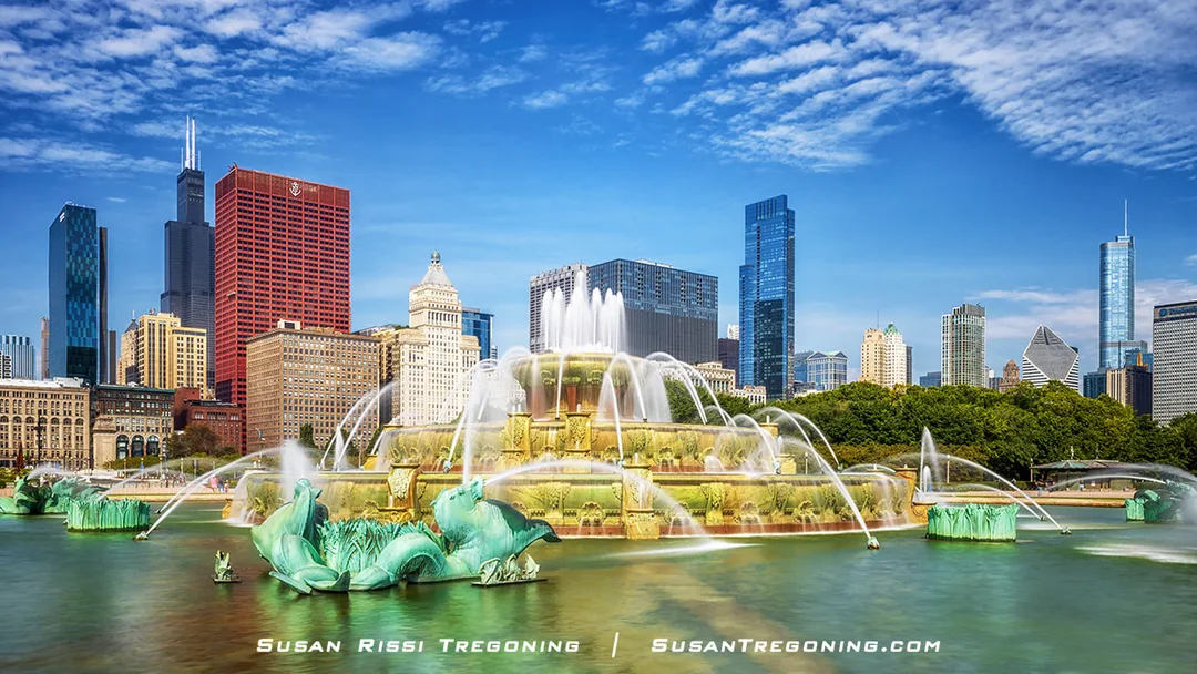 A wide view of Buckingham Fountain in Chicago’s Grant Park with water jets spraying from its tiered basins and sculpted sea horses, set against the downtown skyline including the Willis Tower, the red CNA Center, and other high‑rise buildings under a blue sky with scattered clouds.